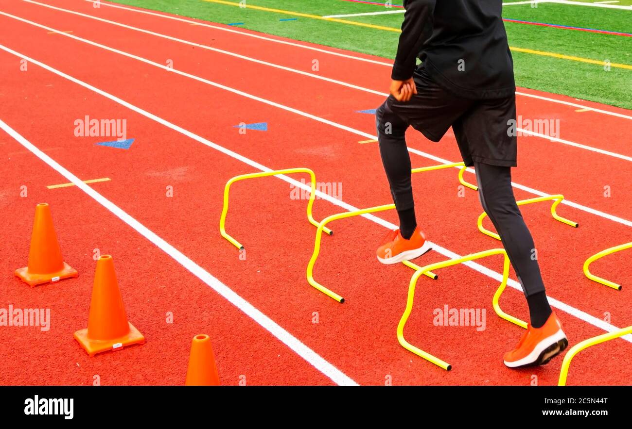 Rear view of a high school track and field athlete is running over yellow hurdles set up in lane on a red track. Stock Photo