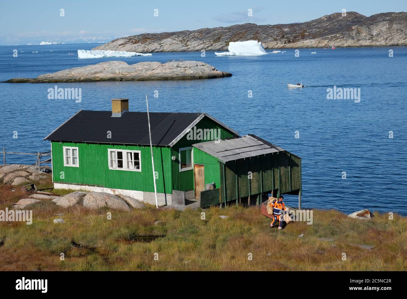 Brightly dressed workman collects the garbage from a small green house perched on the rocks which is dwarfed by huge icebergs floating by. Stock Photo