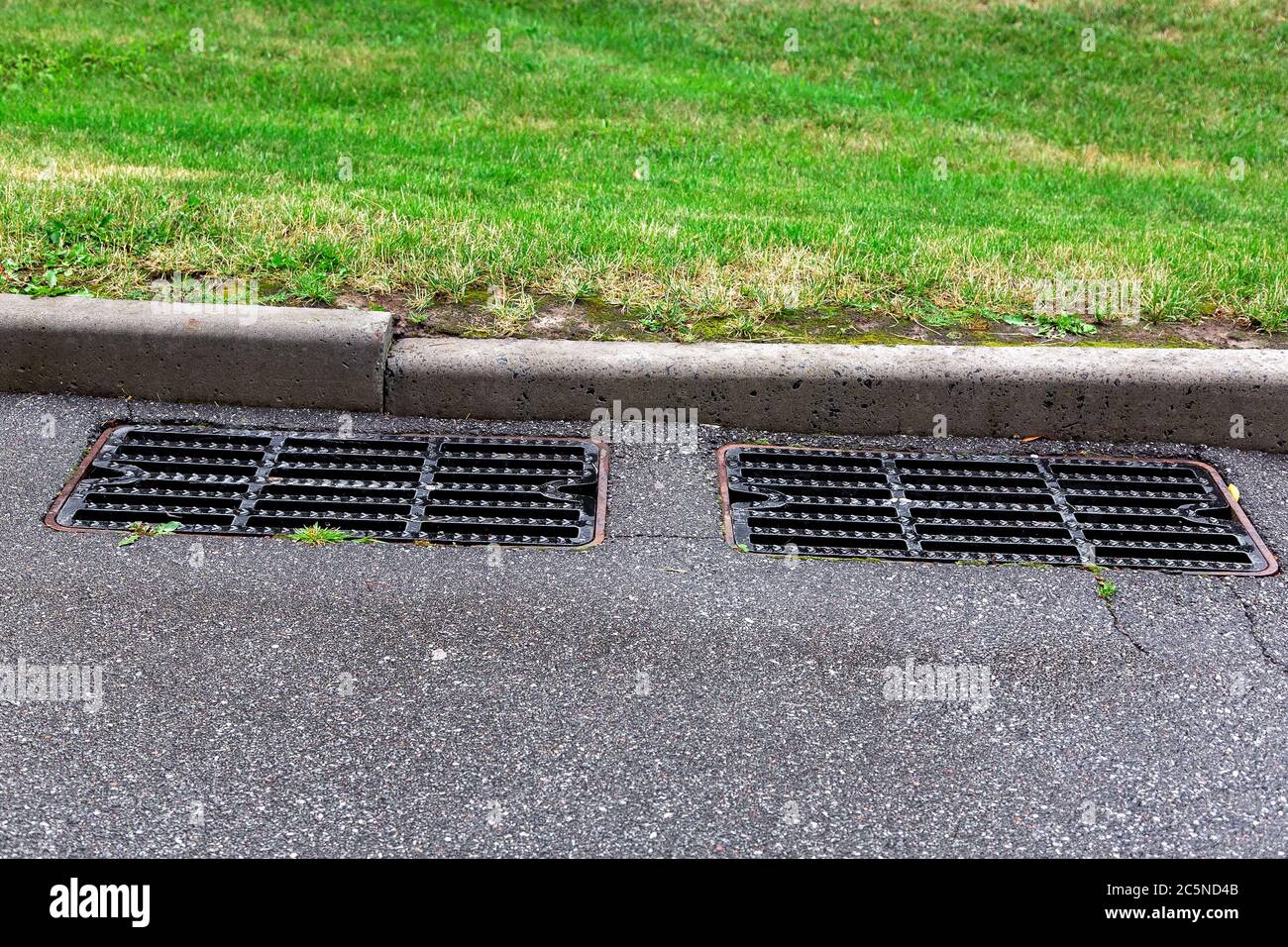 two grilles of a sewer manhole of a storm road system on the slope of an asphalt road with green grass in the background. Stock Photo