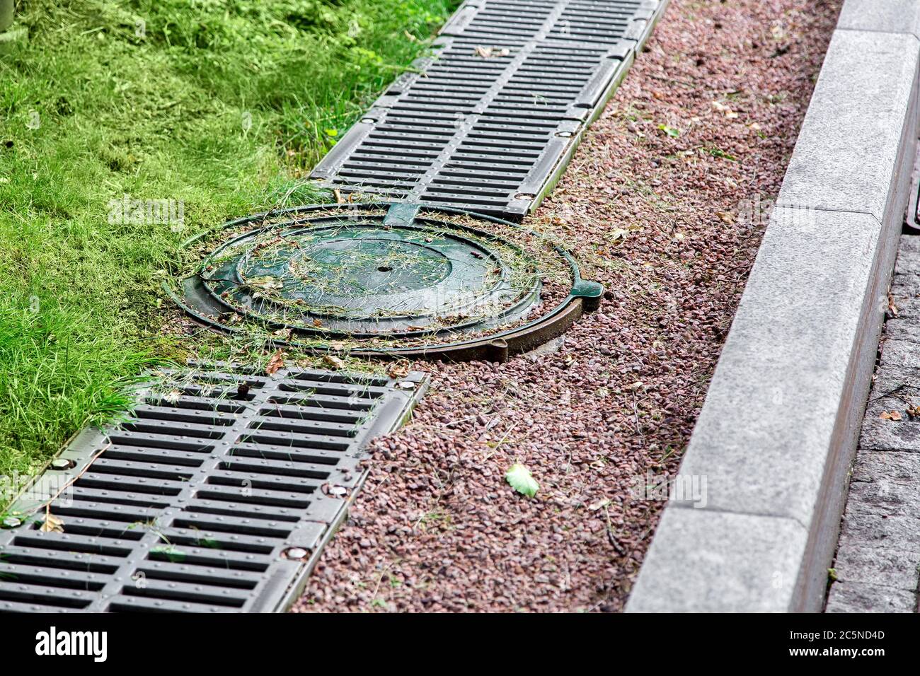 round manhole cover of drainage system on the side of the road with an iron grate and pebbles. Stock Photo
