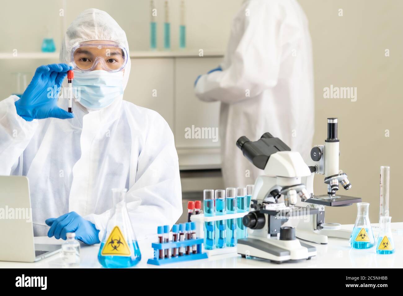Scientists hold blood sample test tube and examine for his research and develop vaccine for coronavirus covid-19 pandemic with his colleague in backgr Stock Photo