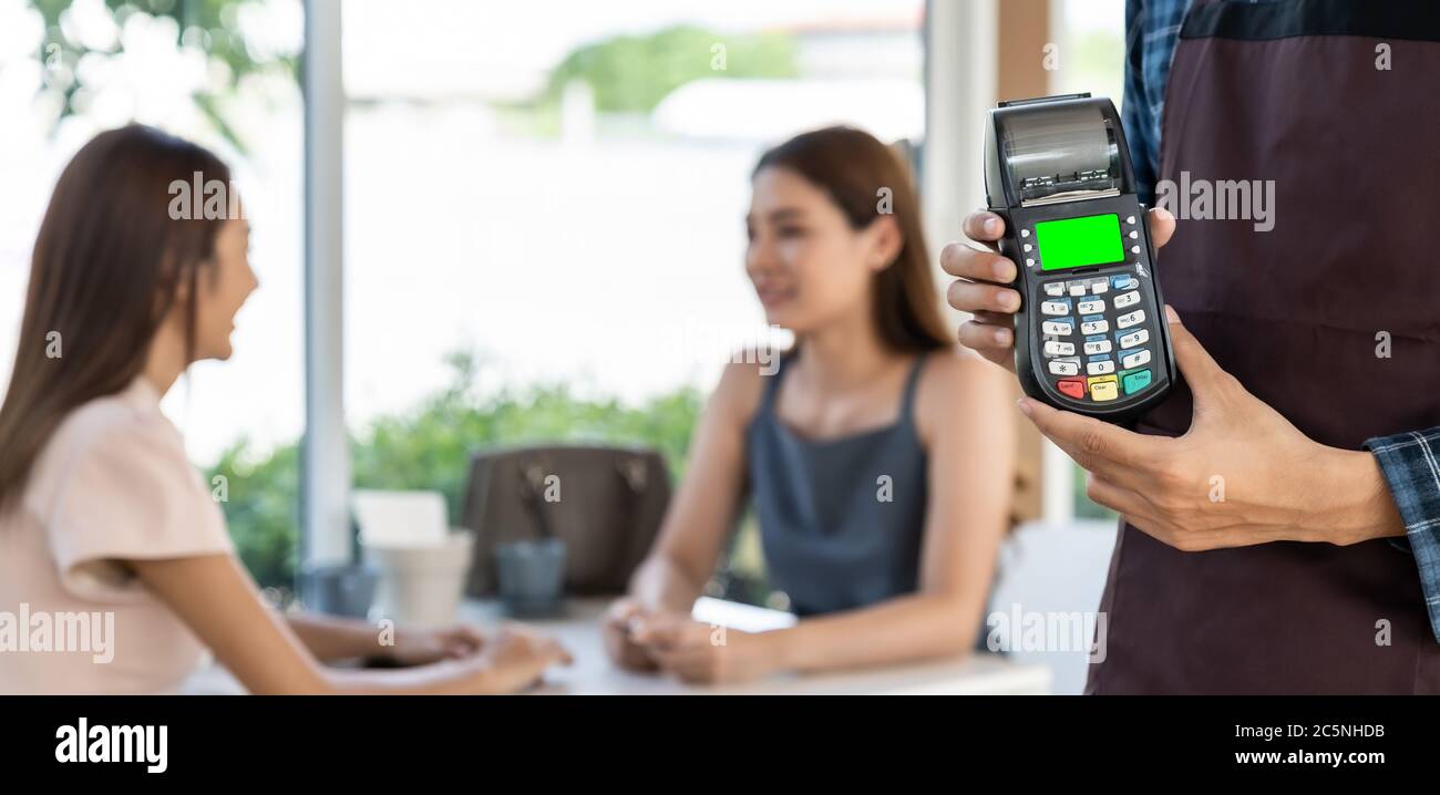 Panoramic close up of waiter hand hold credit card reader for contactless payment with customers in restaurant background. New normal restaurant conta Stock Photo
