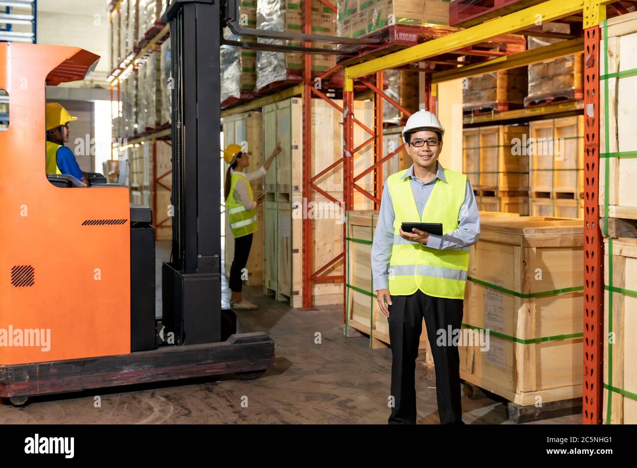 Portrait of Asian warehouse manager hold digital tablet with warehouse worker operate forklift to check inventory in background. Reopening business wa Stock Photo