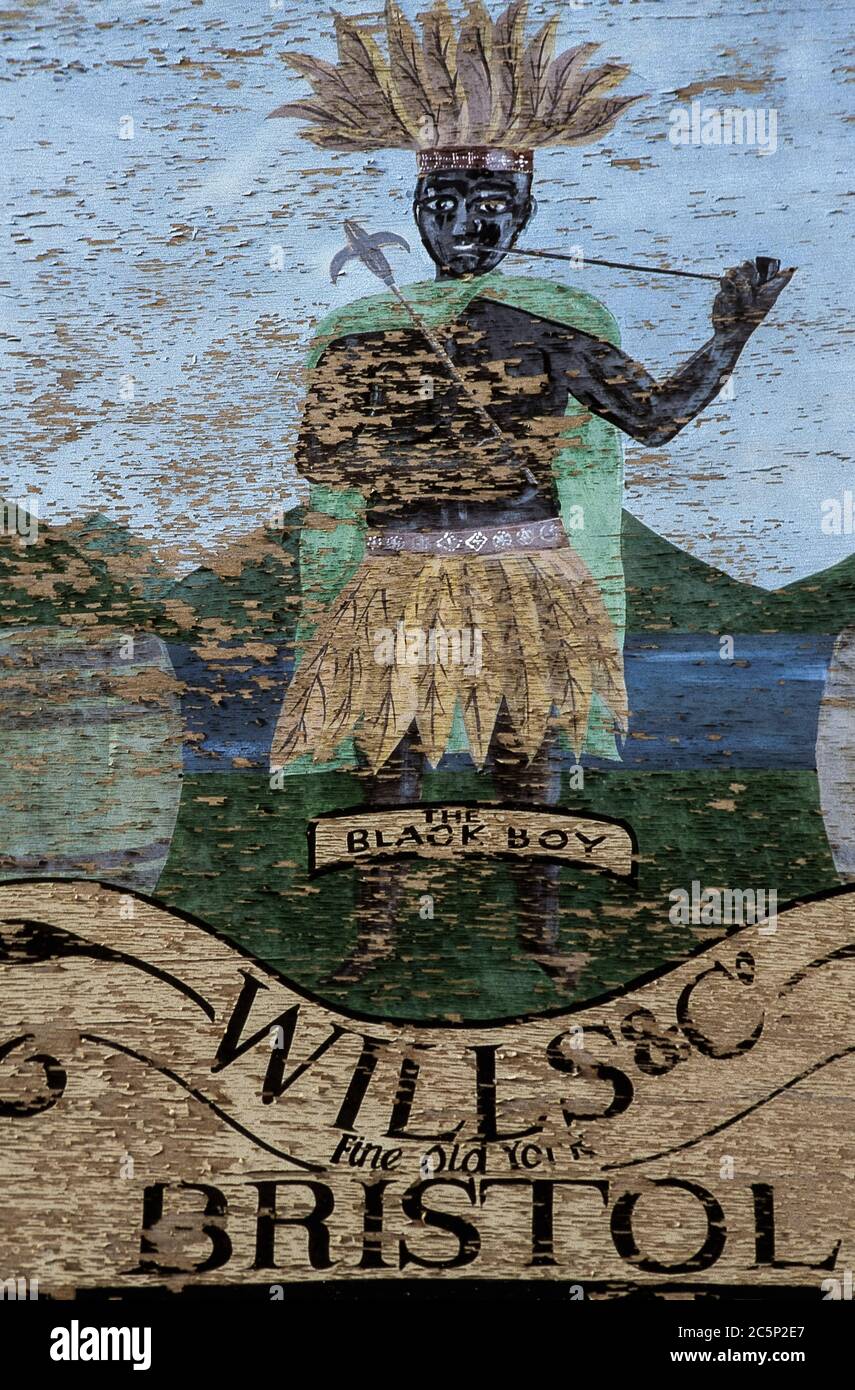 ‘The Black Boy’: a flaking vintage advertisement for tobacco merchants, Wills and Co, hand-painted on a wooden panel - racist and deeply offensive to modern eyes.  The boy is shown smoking a pipe while wearing headgear and tunic or skirt made of tobacco leaves.  Photographed in August 1999 on the façade of tobacconist Marshall’s, also known as The Pipe Shop, at 6 St. Mary’s Street, Ely, Cambridgeshire, England.  Marshall’s closed in 2006 and this vintage panel has long since disappeared. Stock Photo