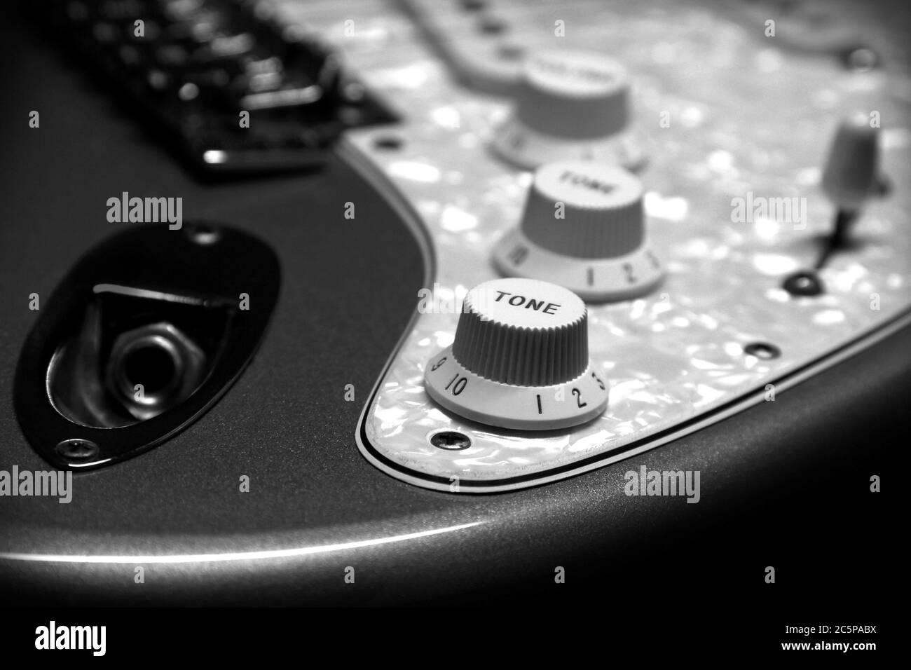 Black and white detail of a Fender Stratocaster electric guitar showing the input jack, tone and volume knobs, selector switch, white pearl pickguard, bridge, and pickups. Stock Photo