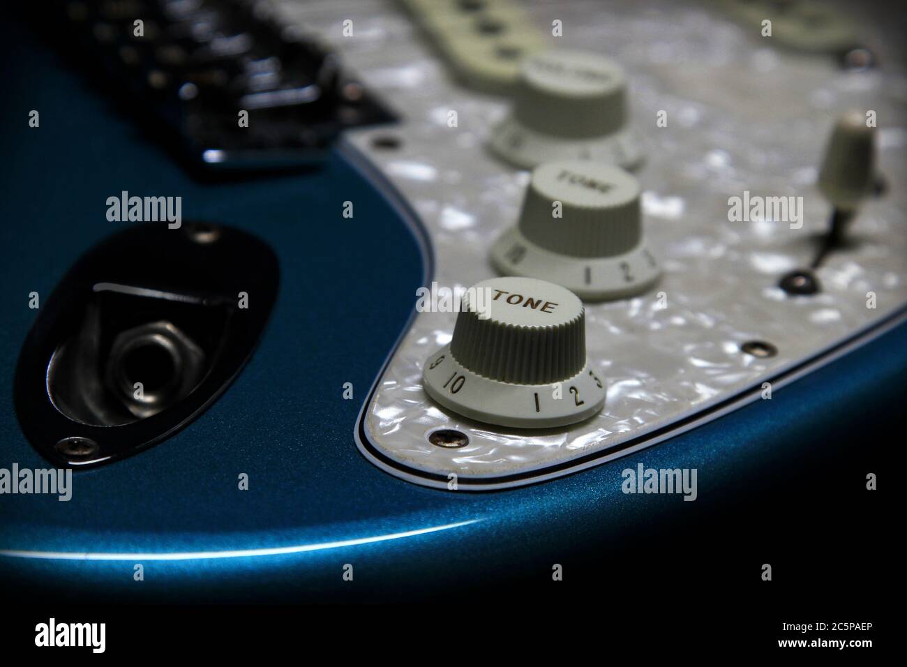 Detail of a Fender Stratocaster electric guitar in Blue Sparkle showing the input jack, tone and volume knobs, selector switch, white pearl pickguard, bridge, and pickups. Stock Photo