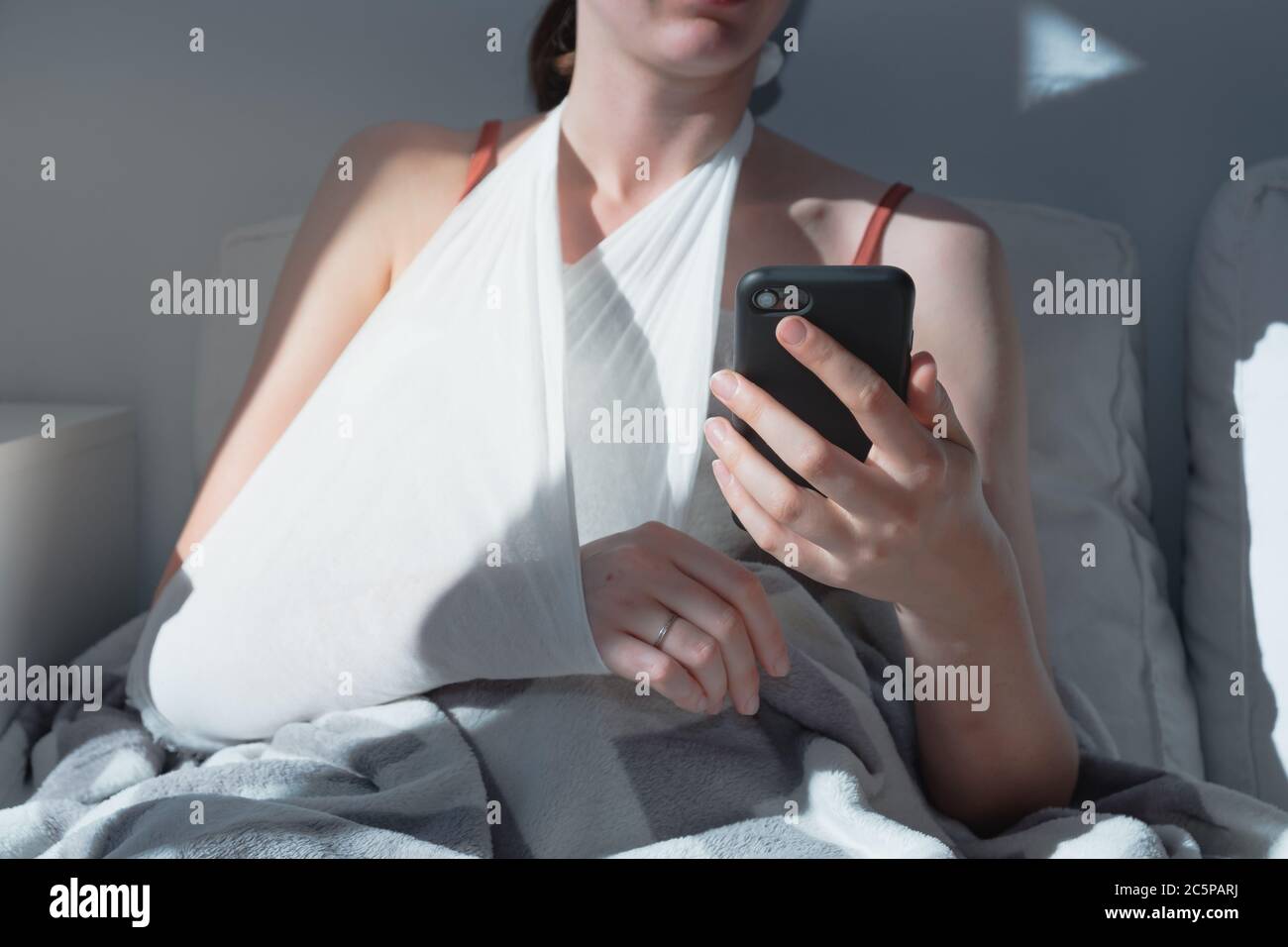 Wounded arm in bondage, woman rests in  bed, body parts. Caucasian female adult in bright sun-lit room holds a phone, concept of self treatment, medic Stock Photo