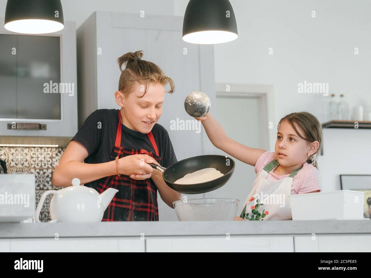 Sister and Brother dressed aprons making a homemade pancakes on the home kitchen. Girl poring a liquid dough on the hot pan. Kids Home cooking concept Stock Photo