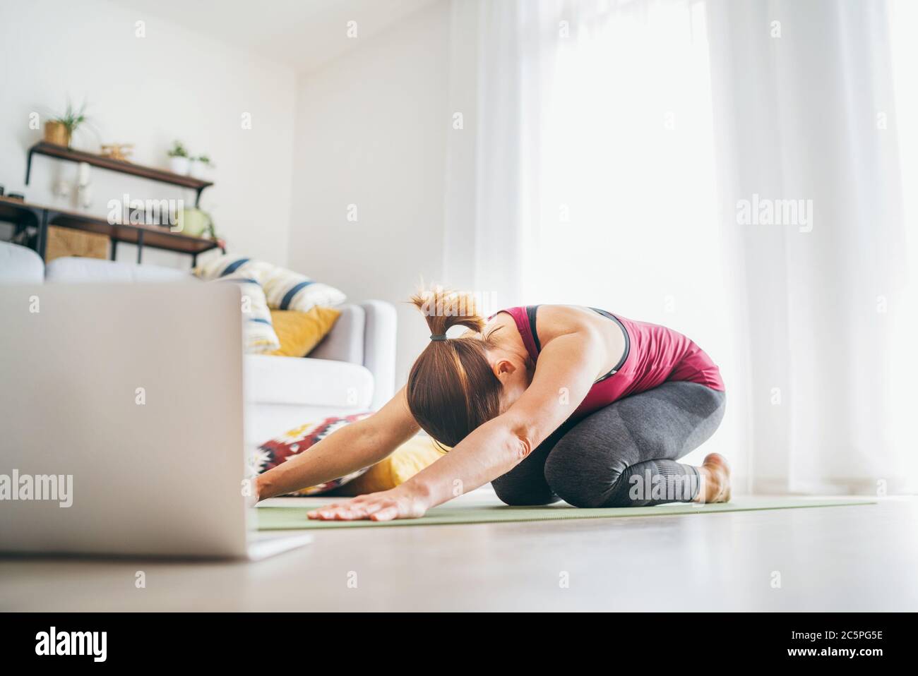 Fit sporty healthy woman on mat in Child’s Balasana gentle resting yoga pose, doing breathing exercises, watching online yoga class on laptop computer Stock Photo
