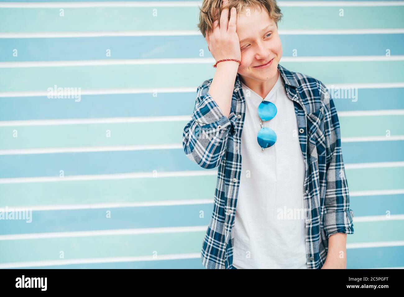 Fashion portrait of caucasian blue-eyed blonde hair 12 year old teenager boy dressed t-shirt and checkered shirt with sunglasses cramped because of sh Stock Photo