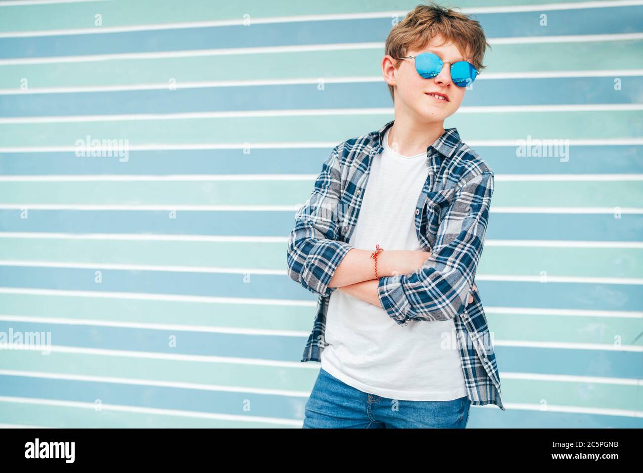 Fashion portrait of caucasian blonde hair 12 year old teenager boy dressed t-shirt and checkered shirt in blue sunglasses posing on turquoise blue bac Stock Photo