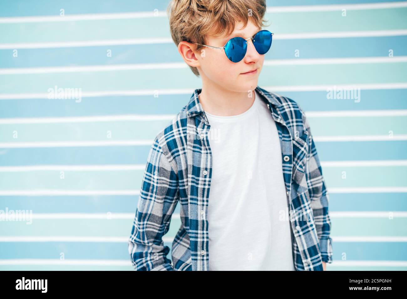 Fashion portrait of caucasian blonde hair 12 year old teenager boy dressed white t-shirt with checkered shirt in blue sunglasses with turquoise blue b Stock Photo