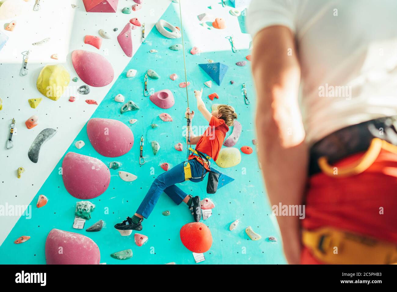 Father and teenager son at indoor climbing wall hall. Boy is hanging on the rope using a climbing harness and daddy belaying him on the floor using a Stock Photo