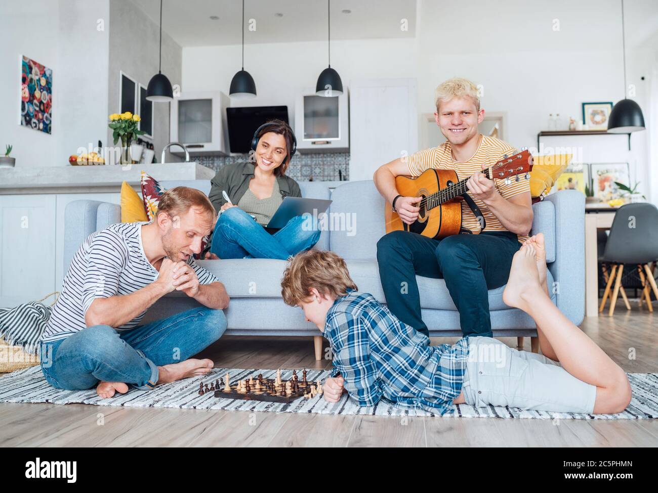 Family at living room together. Father playing chess with little son, mother learning online using laptop, older son playing acoustic guitar and singi Stock Photo