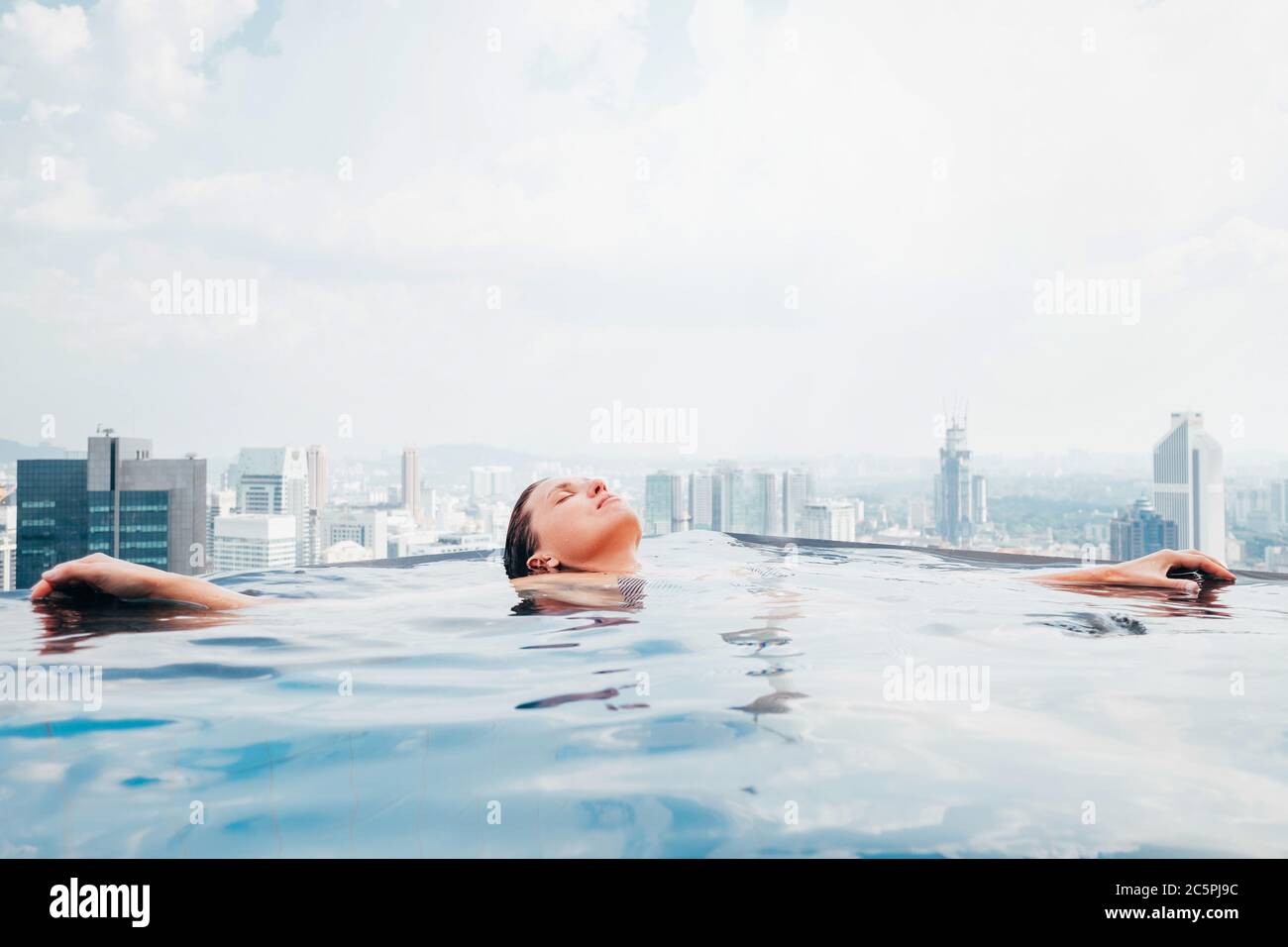 Young female relaxing in top roof infinity swimming pool with city view to the modern business center downtown with a skyscrapers and towers. Luxury v Stock Photo