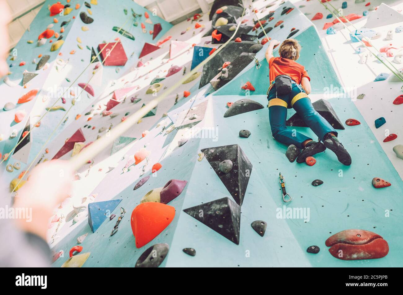 Teenager boy at indoor climbing wall hall. Boy is climbing using a top rope and climbing harness and somebody belaying him from floor. Active teenager Stock Photo
