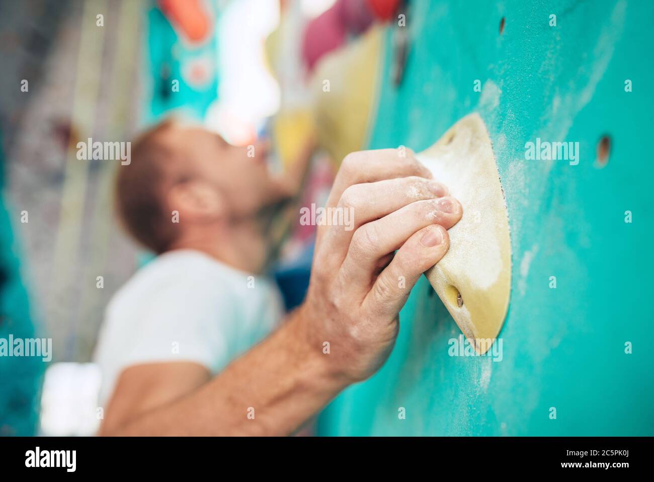 Close-up shot of chalk (magnesium) covered man's palm clinging the indoor climbing grip element while man climbing up. Unfocused climbers face on back Stock Photo