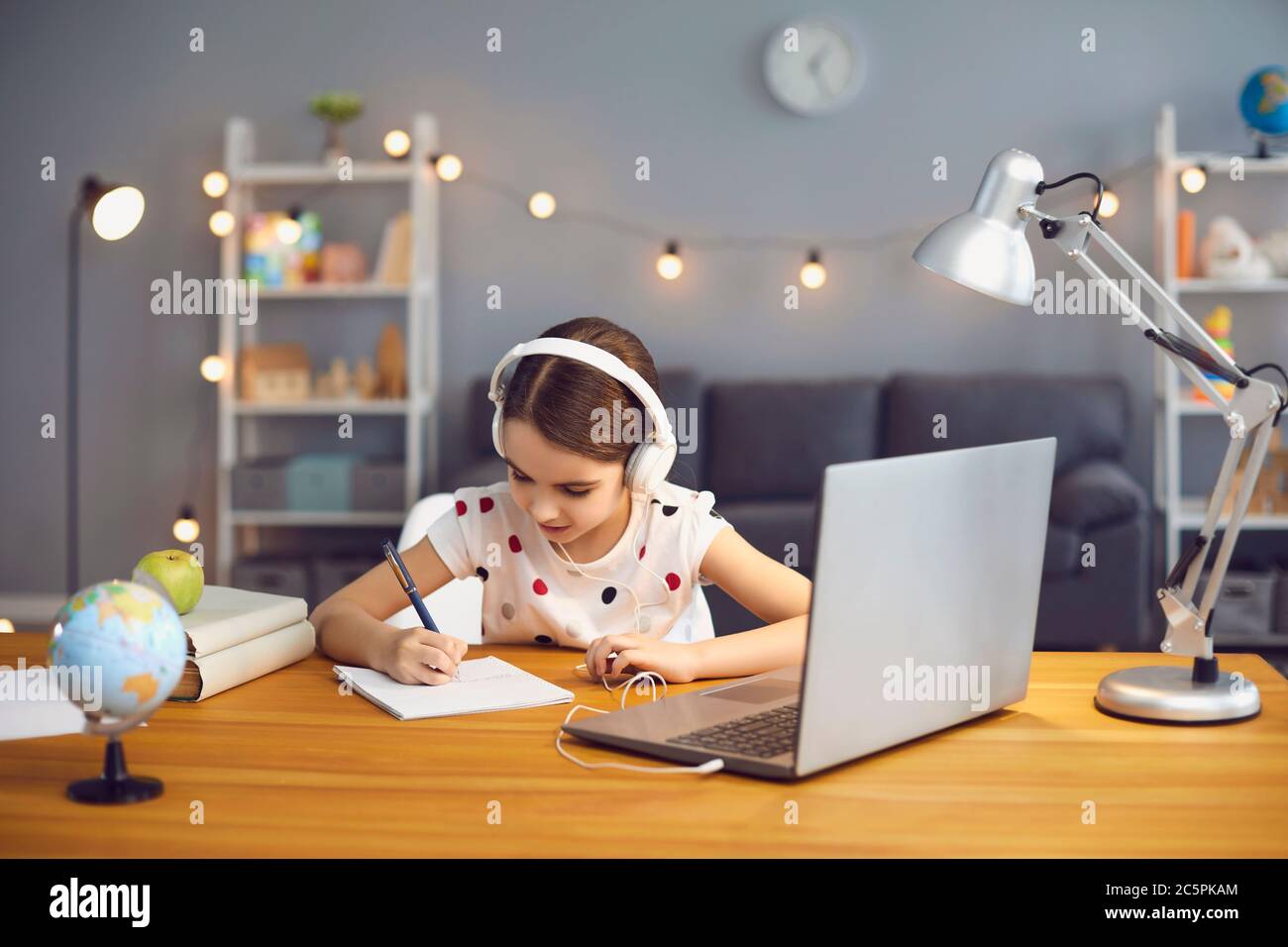 Serious little girl with headphones and laptop studying online at home, writing down notes in her copybook Stock Photo