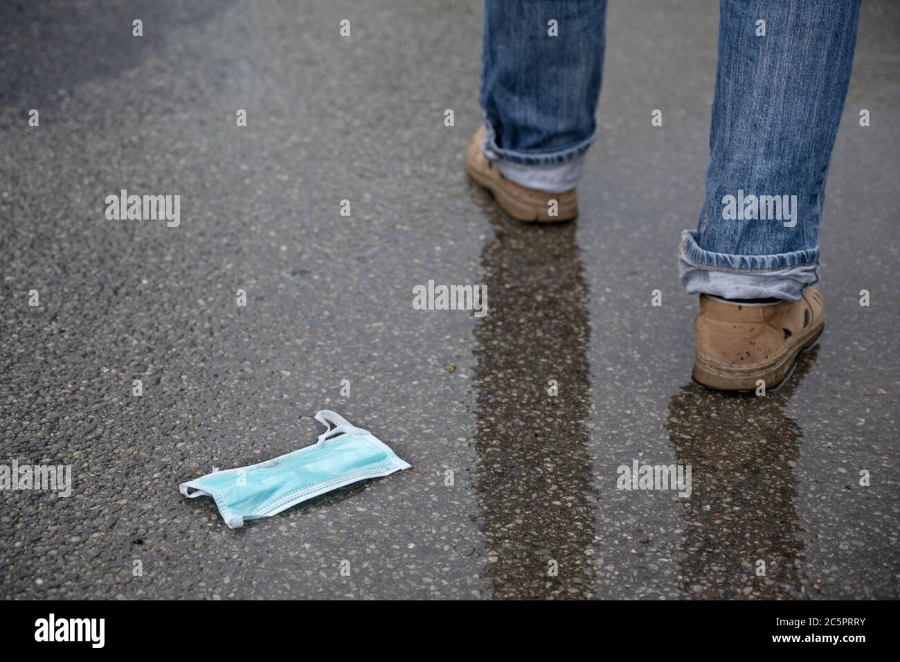 Medical face mask discarded on the wet asphalt, man's legs walk  away, reckless behavior against the risk of coronavirus infection after the lockdown, Stock Photo
