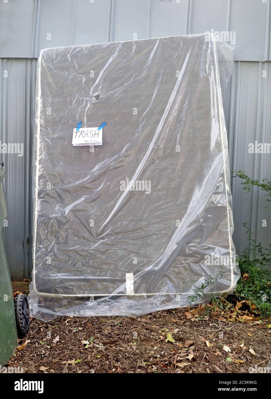 The proper way to dispose of used mattress. Abandoned mattress leaning against garage in alley wrapped in plastic with TRASH note attached. Stock Photo