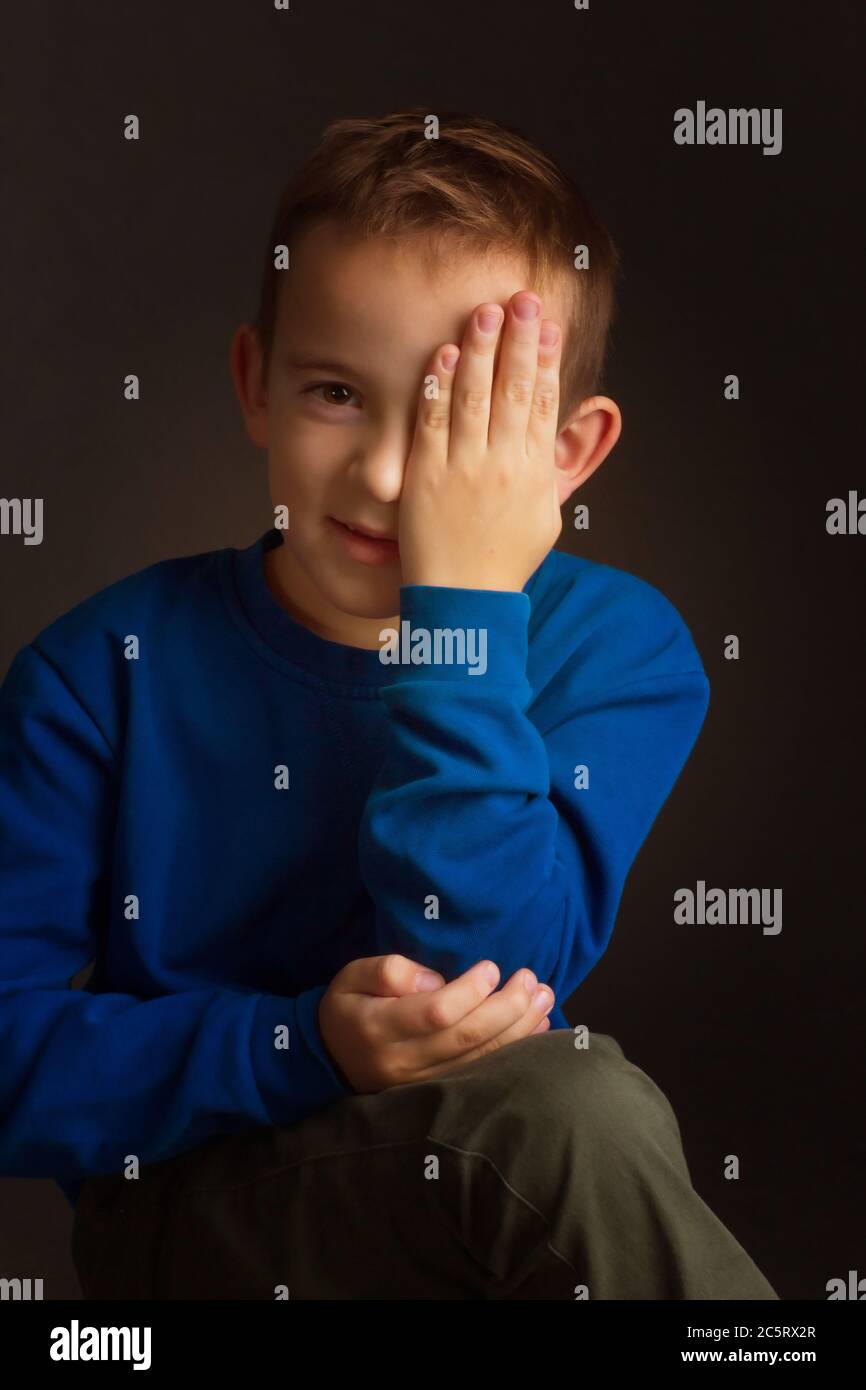 portrait of a dark-haired boy covering his eye with his hand on a black ...