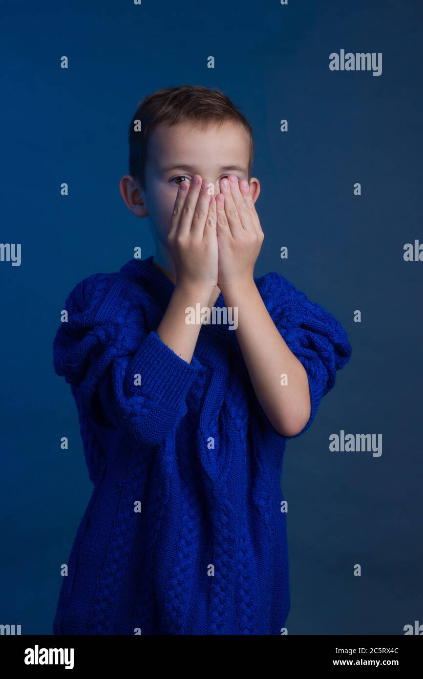 Studio portrait of unhappy boy in a blue classic sweater on a blue background. hiding peeking out from behind his hands Stock Photo