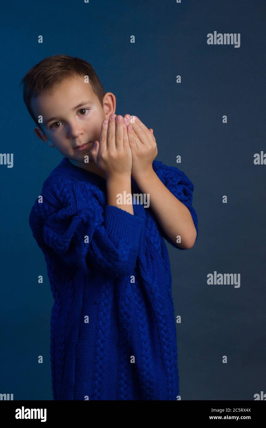 Studio portrait of unhappy boy in a blue classic sweater on a blue background. hiding peeking out from behind his hands Stock Photo
