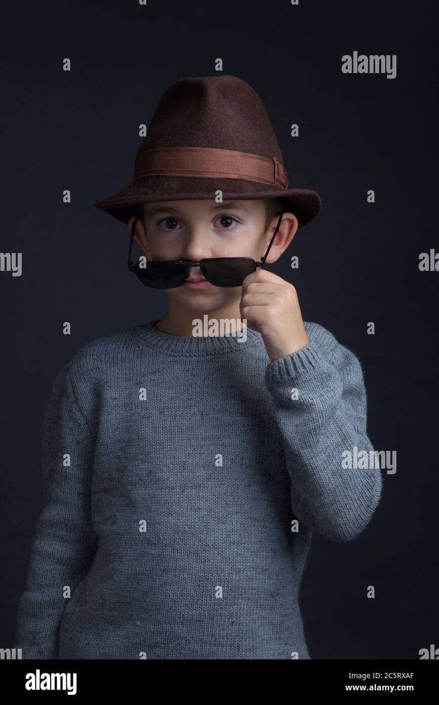 Studio portrait of a boy in a grey sweater, brown hat and holds the glasses at nose level with his hand, on black background Stock Photo
