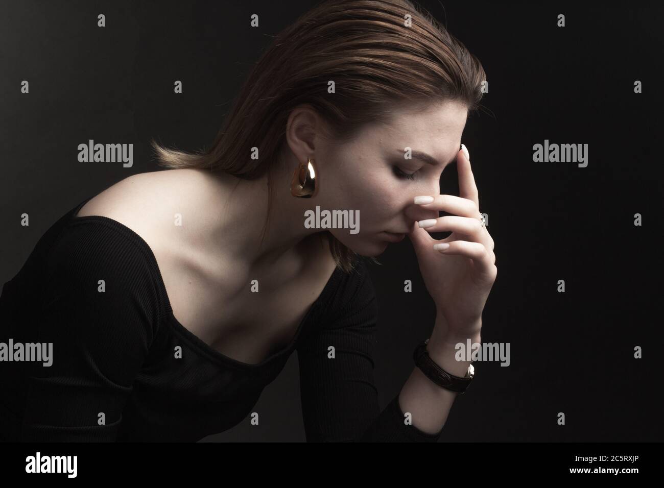 Portrait in the photo Studio of a brown-haired girl, on a black background in black clothes with open shoulders. Short hair. turned in profile, hand to touch the face and nose. Pensive and sad expression Stock Photo