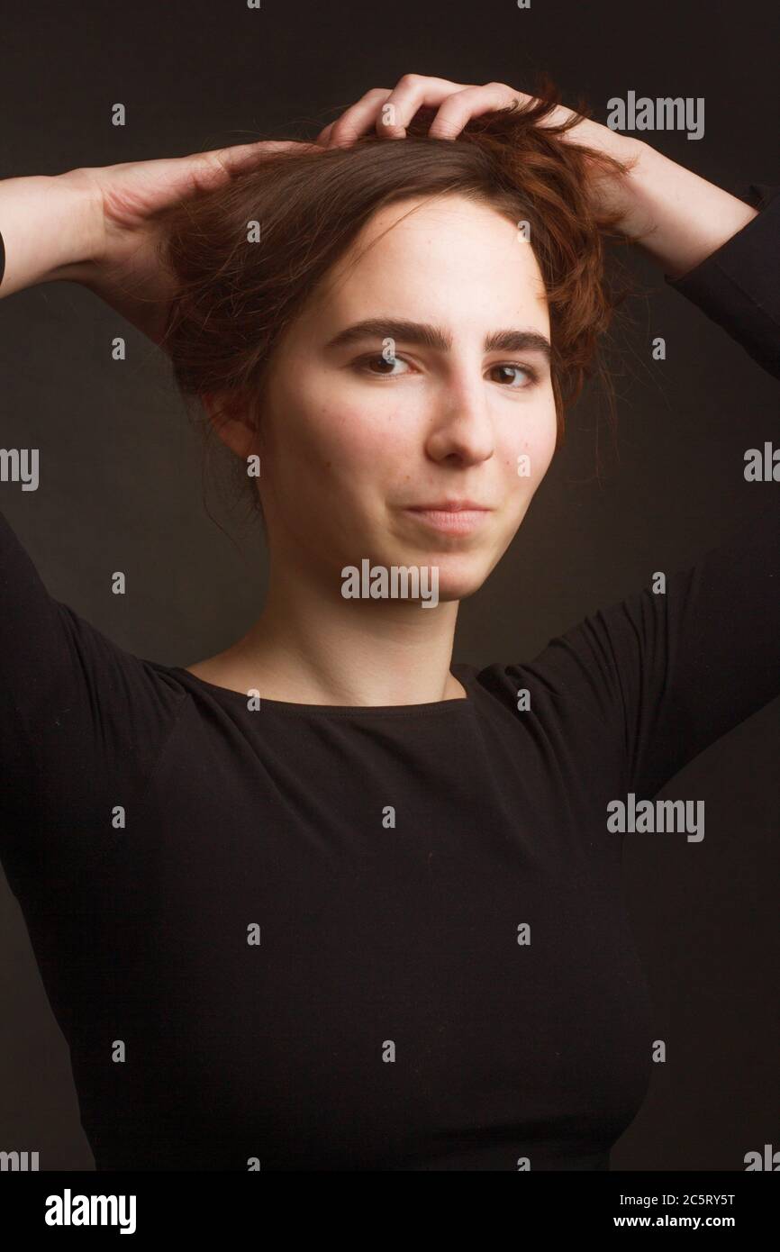 The brown-haired girl in dark clothes, a portrait on a dark gray Studio background. Warm toning. Keeps hands above the head, the hair, fix the hair Stock Photo