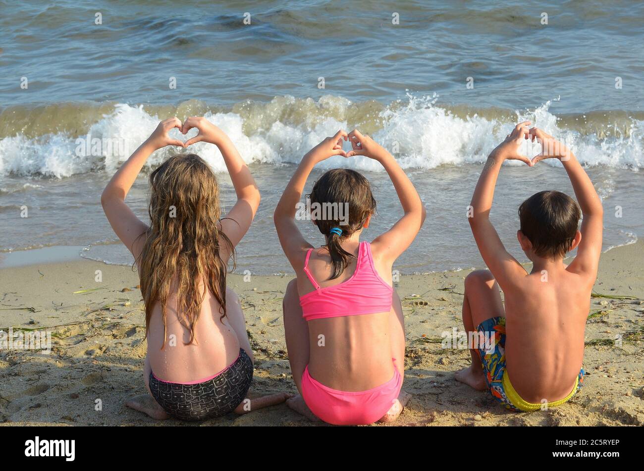 two girls and a boy sitting on a sandy beach in swimming suits, facing the  sea, making hearts with their hands above their heads Stock Photo - Alamy