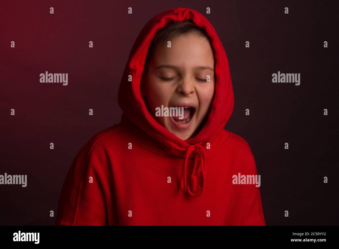 Studio portrait of a dark-haired boy, on a maroon background, in a red hoodie jacket. Positive emotion, smile on your face, yawns Stock Photo