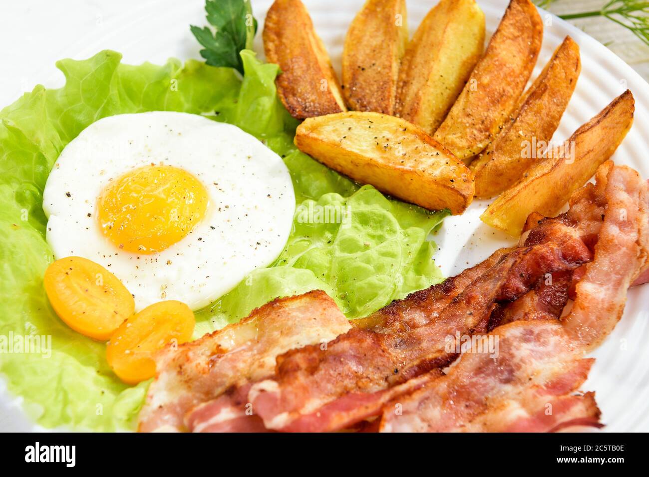French fries and egg with bacon on top in a white bowl, on a large table with greens. Stock Photo