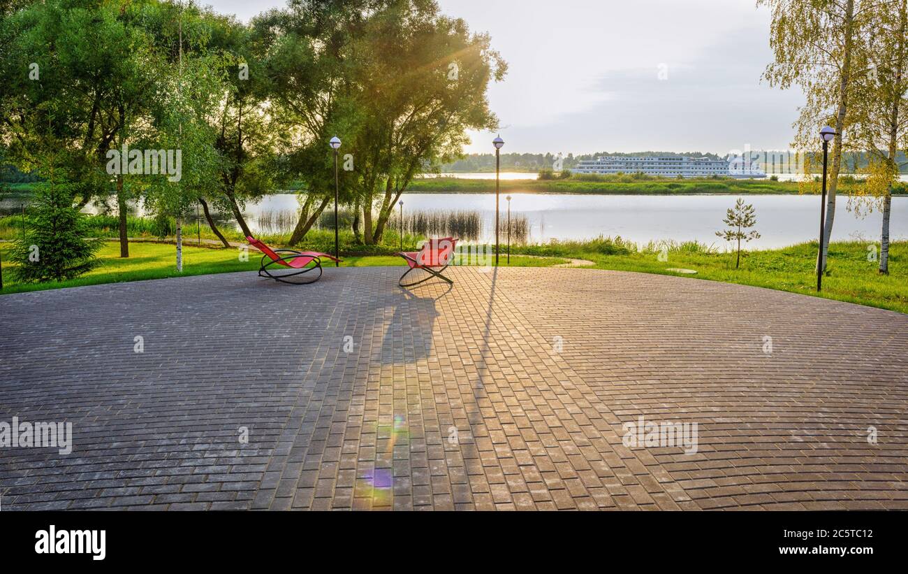 Two red armchairs, standing by the bank of the river, the sun is shining, and a white, large steamer is sailing along the river. Stock Photo