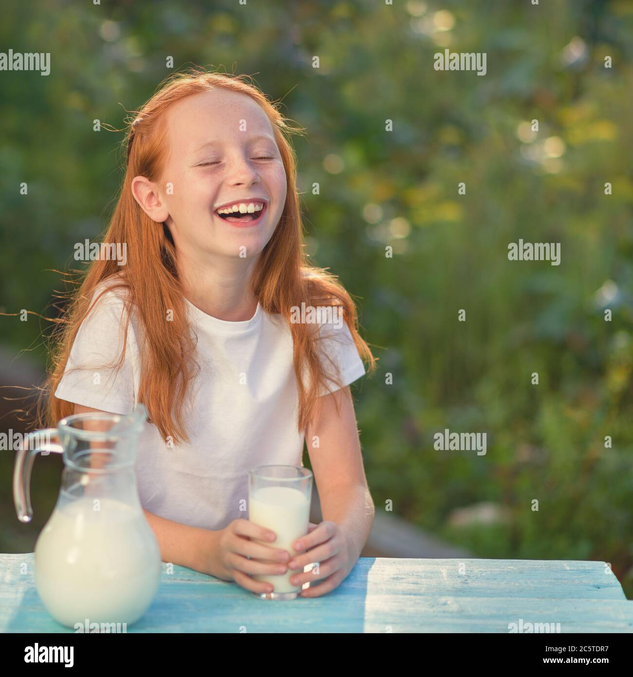 Milk is fresh from the cow - a beautiful girl with dairy products. Girl holds glass with fresh milk. Happy redhead child in summer. Closeup of little Stock Photo