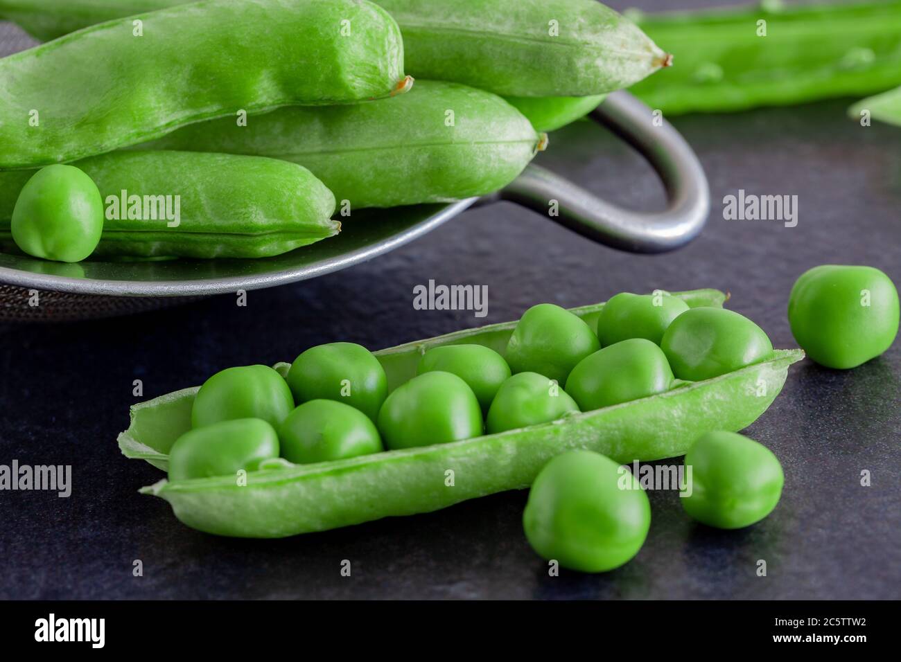 open pea pods on a dark table with a sieve in the background Stock Photo