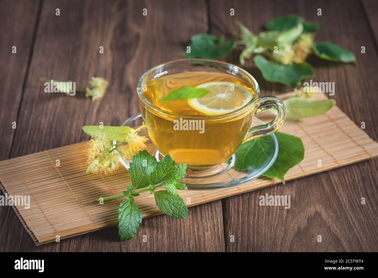 A cup of medicinal, lime tea, standing on a wooden table, surrounded by fragrant linden flowers, in the rays of sunlight. Stock Photo