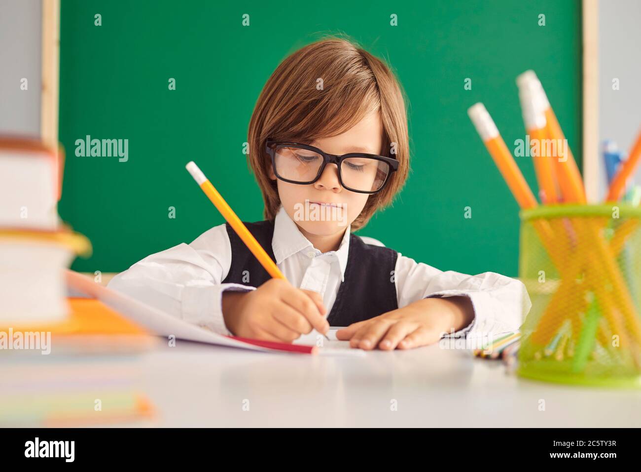 Schooling. A smart schoolboy with glasses writes a teacher assignment in a notebook at a table in the classroom. Stock Photo