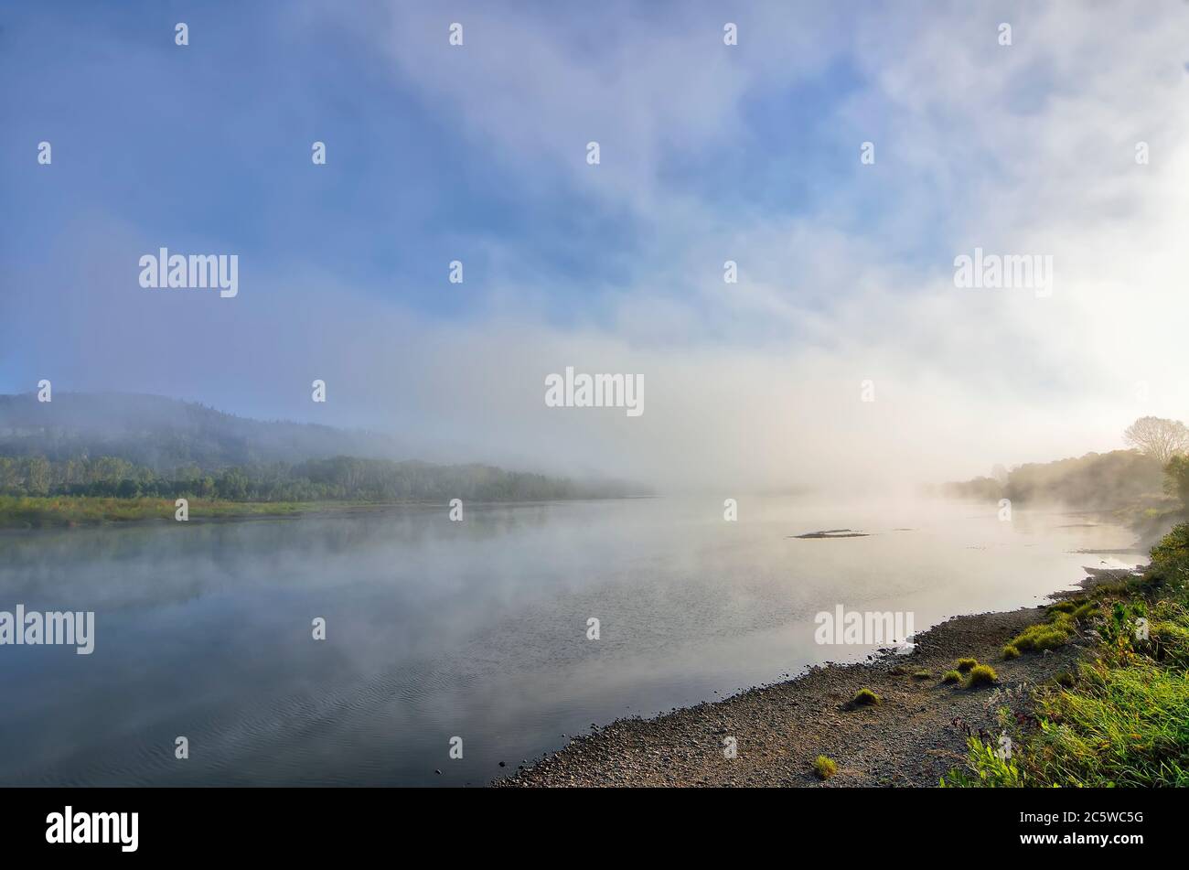 Early foggy morning over the river - beautiful summer landscape. Thick fog over the water surface, reflection of hill with forest covered on the bank Stock Photo