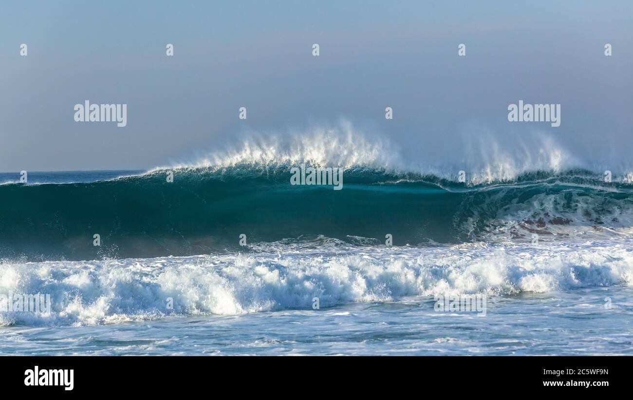 Ocean wave feathering upright wall of blue water flaring and crashing on shallow reef sandbars closeup panoramic photo. Stock Photo