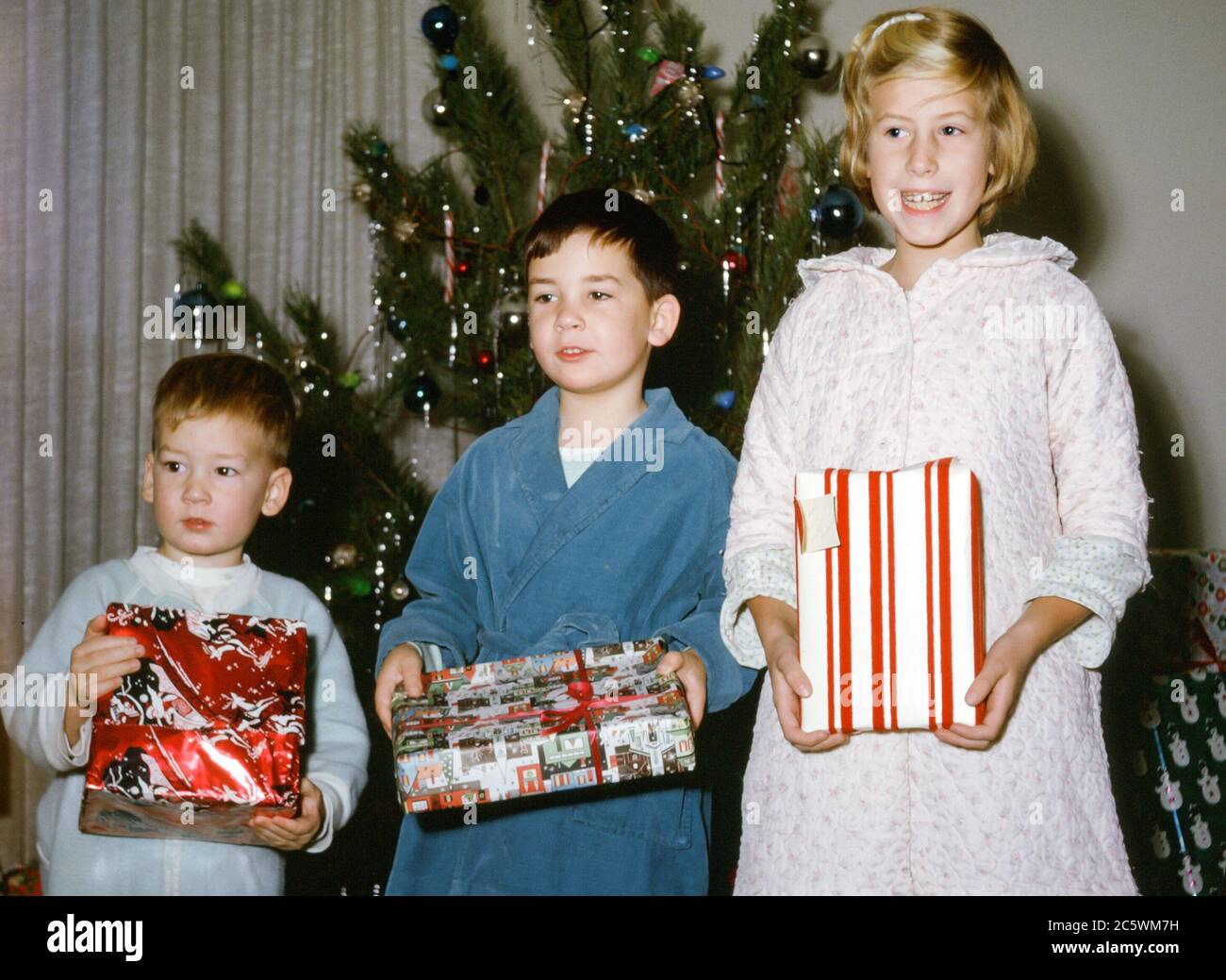 Three young children, siblings comprising a 7 year old girl, 5 year old boy and 3 year old boy, in pyjamas and dressing gowns holding Christmas presents in front of a Christmas tree on Christmas morning in USA in the  1960s Stock Photo