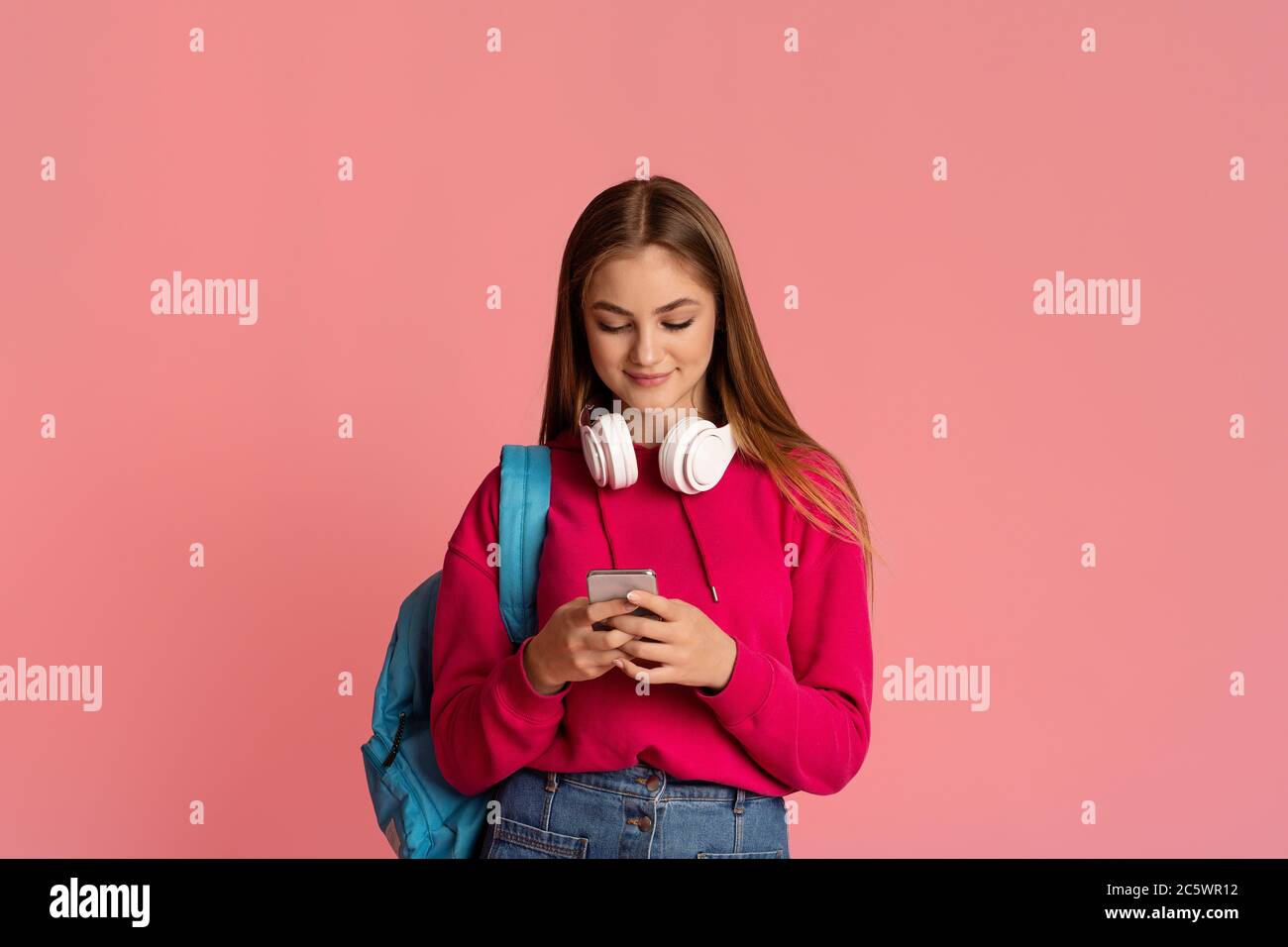 Modern teenagers. Smiling girl with headphones and backpack tapping on smartphone Stock Photo