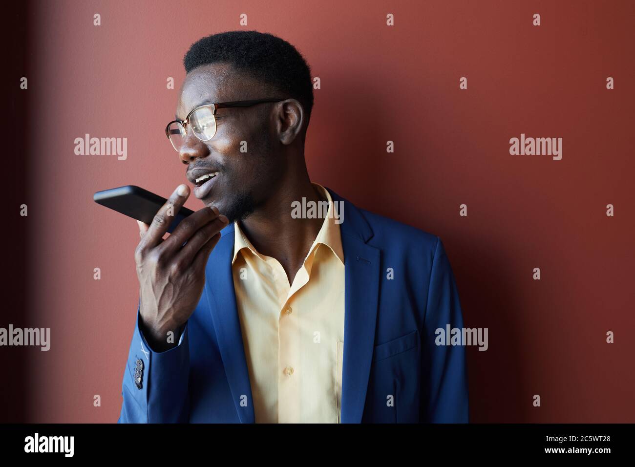 Portrait of elegant African-American man recording voice message via smartphone and wearing glasses while posing against maroon background, copy space Stock Photo