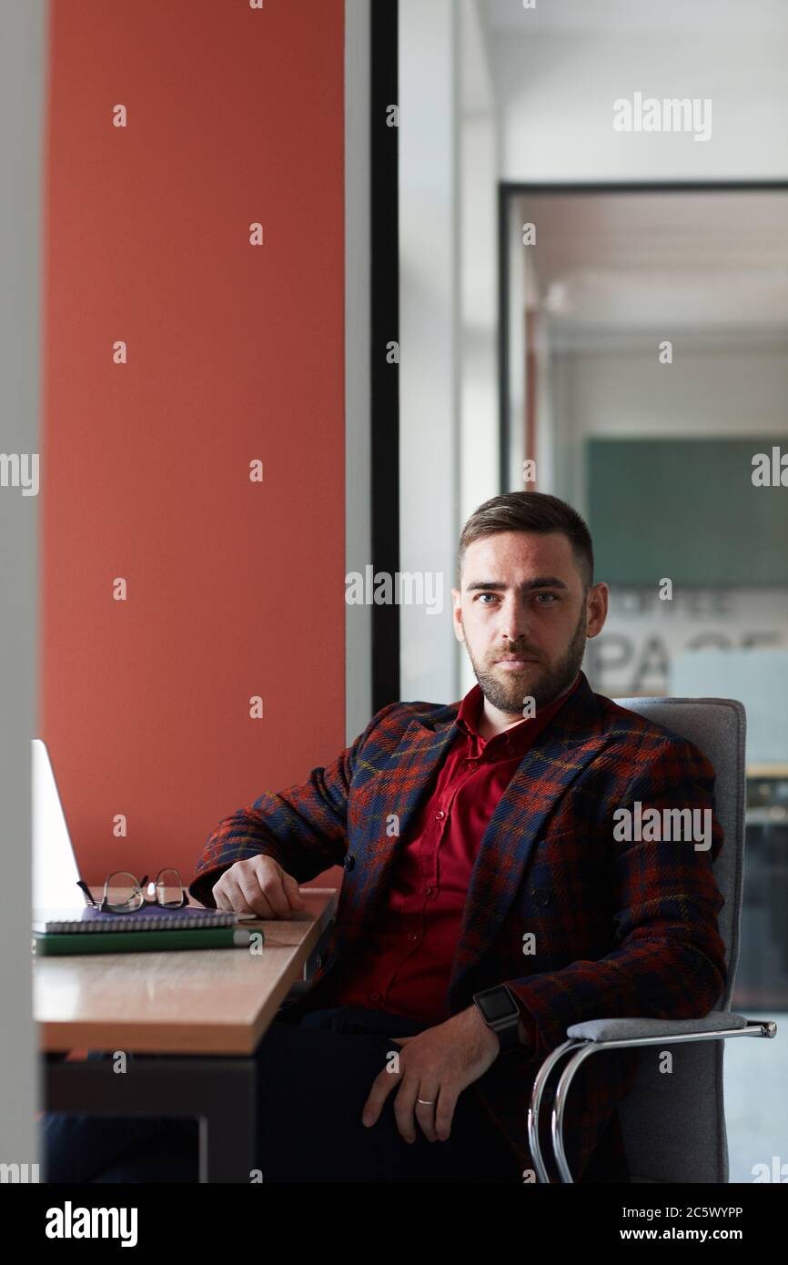 Vertical portrait of elegant bearded businessman looking at camera while sitting at workplace and posing confidently against red wall in office, copy Stock Photo