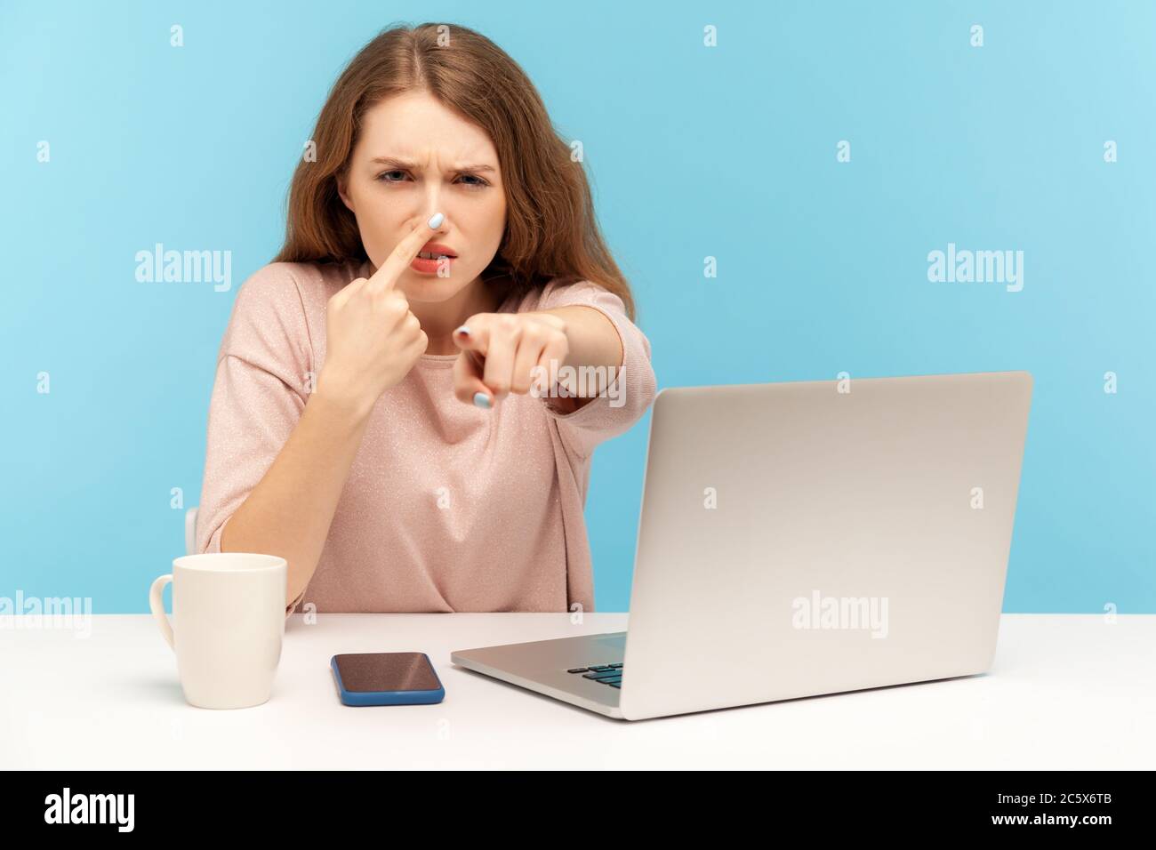 You are liar! Angry young woman employee sitting at workplace, touching nose and pointing to camera, blaming liar in deception, suspecting falsehood. Stock Photo