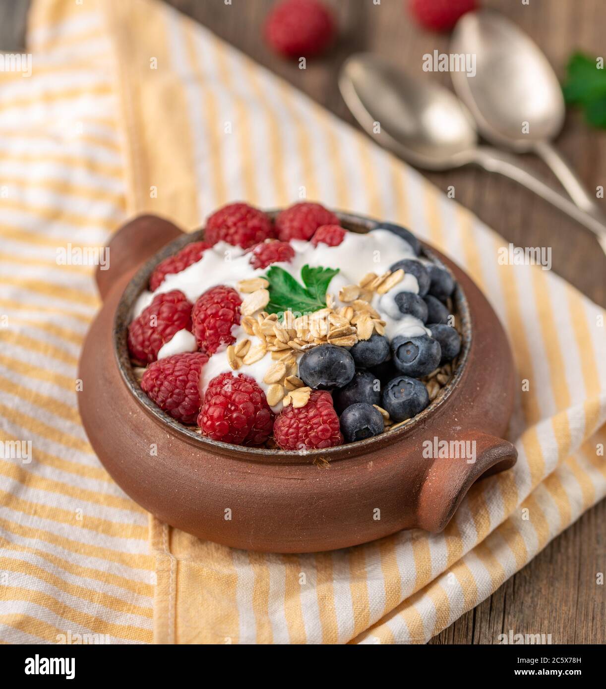 Granola with white yogurt with raspberries and blueberries in ceramic bowl on stripped napkin. Stock Photo