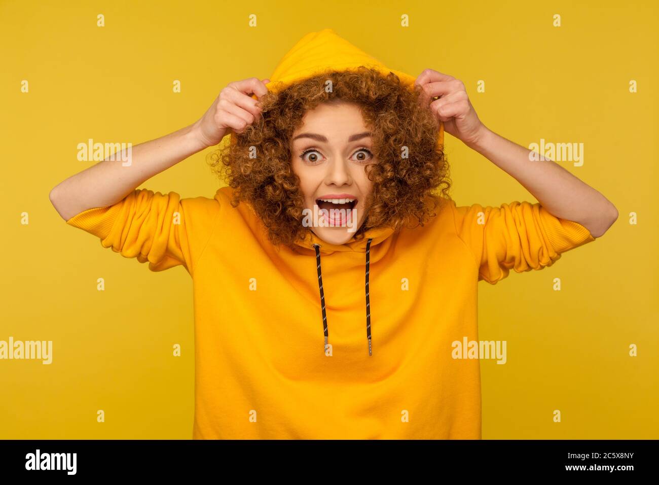 Portrait of funny amazed girl with curly hair wearing urban style hoodie, putting on hood and looking surprised at camera, youth sport style female fa Stock Photo