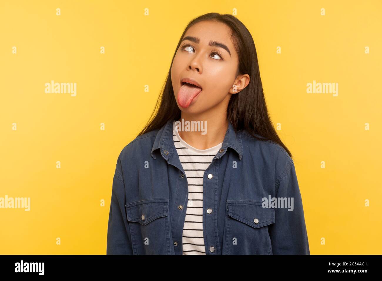 Portrait of funny dumb crazy girl in denim shirt showing tongue out and looking cross-eyed with silly brainless expression, aping fooling around. indo Stock Photo