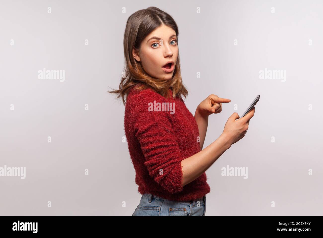 Side view, surprised brunette girl in shaggy sweater pointing at mobile phone and looking at camera with astonished expression, online communication. Stock Photo