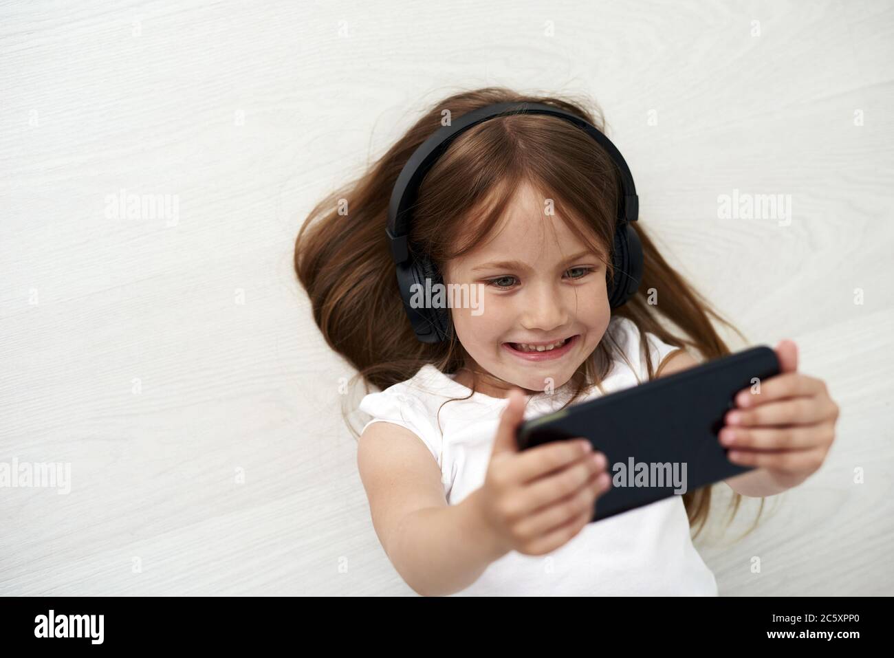 The little is laying on the floor. The girl is holding phone. The little girl is playing phone on the background. Stock Photo